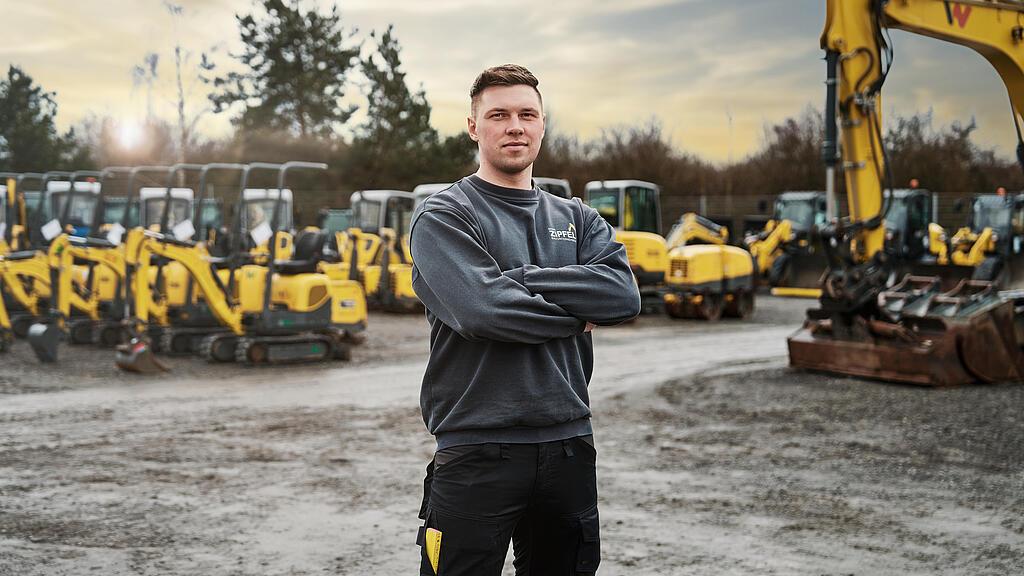 A man stands in front of several yellow mini excavators and construction equipment in a construction machinery yard.