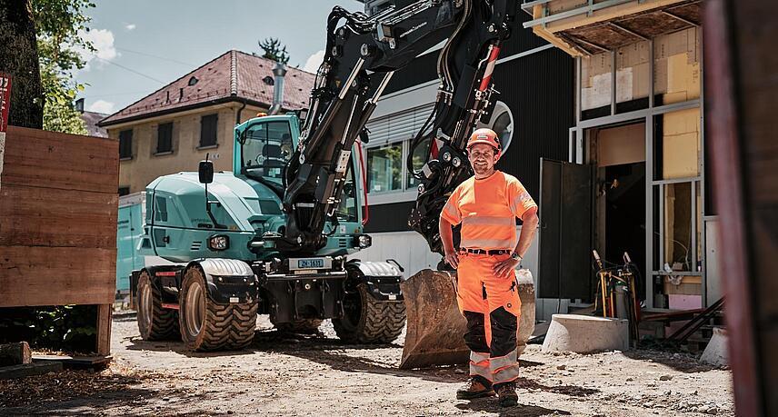 Operator w odzieży roboczej o wysokiej widoczności stojący obok koparki kołowej Wacker Neuson EW100 na miejskim placu budowy.