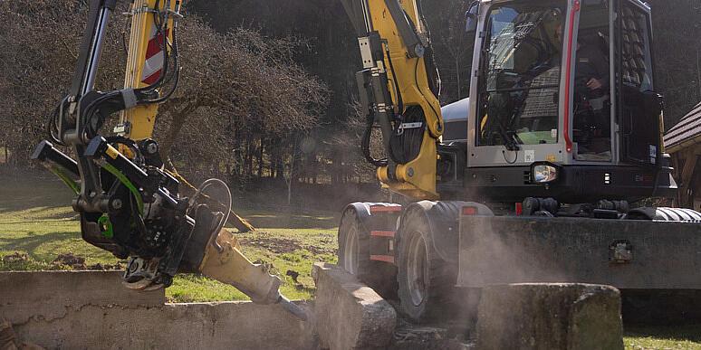 Koparka kołowa Wacker Neuson EW100 obsługująca rozdrabniacz pni na leśnej polanie, rozdrabniająca pień drzewa i wyrzucająca w powietrze wióry drzewne, a w tle ułożone w stosy kłody i las.
