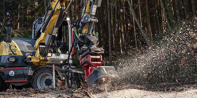 Koparka kołowa Wacker Neuson EW100 obsługująca rozdrabniacz pni na leśnej polanie, rozdrabniająca pień drzewa i wyrzucająca w powietrze wióry drzewne, a w tle ułożone w stosy kłody i las.