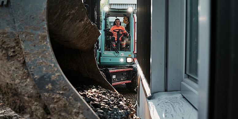 Koparka Wacker Neuson EW100 transportująca kruszywo za pomocą łyżki w wąskim obszarze budowlanym przy budynku.