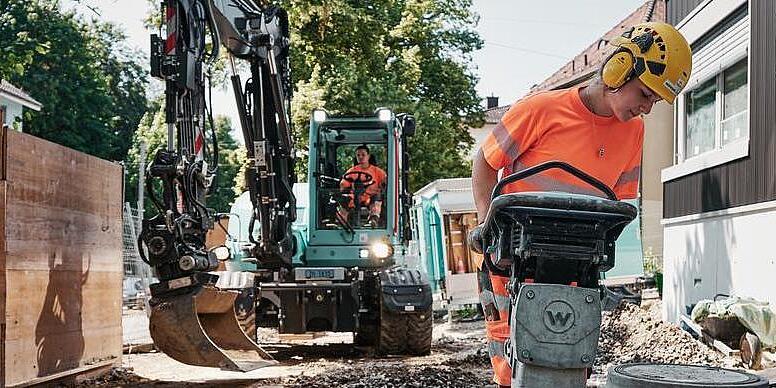 Pracownik budowlany obsługujący ubijak firmy Wacker Neuson, podczas gdy w tle pracuje koparka EW100.