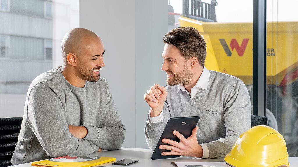 Pracownik firmy Wacker Neuson siedzi przy biurku i pokazuje coś klientowi na tablecie.