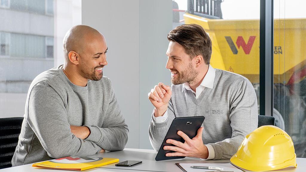 Pracownik firmy Wacker Neuson siedzi przy biurku i pokazuje coś klientowi na tablecie.