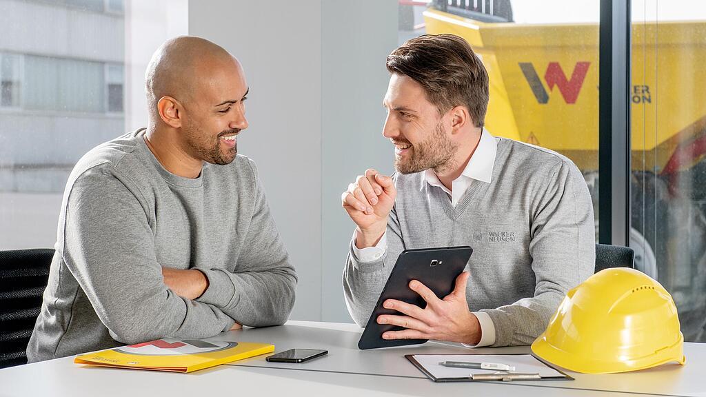 Pracownik firmy Wacker Neuson siedzi przy biurku i pokazuje coś klientowi na tablecie.