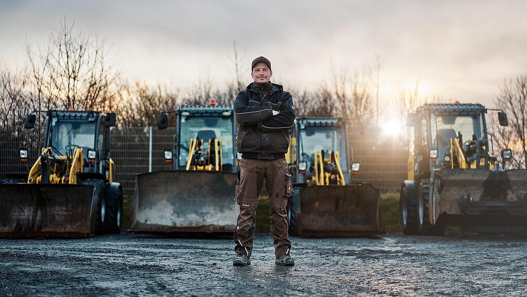 Ein Mann mit verschränkten Armen steht auf einem Hof vor vier gelben Radladern.