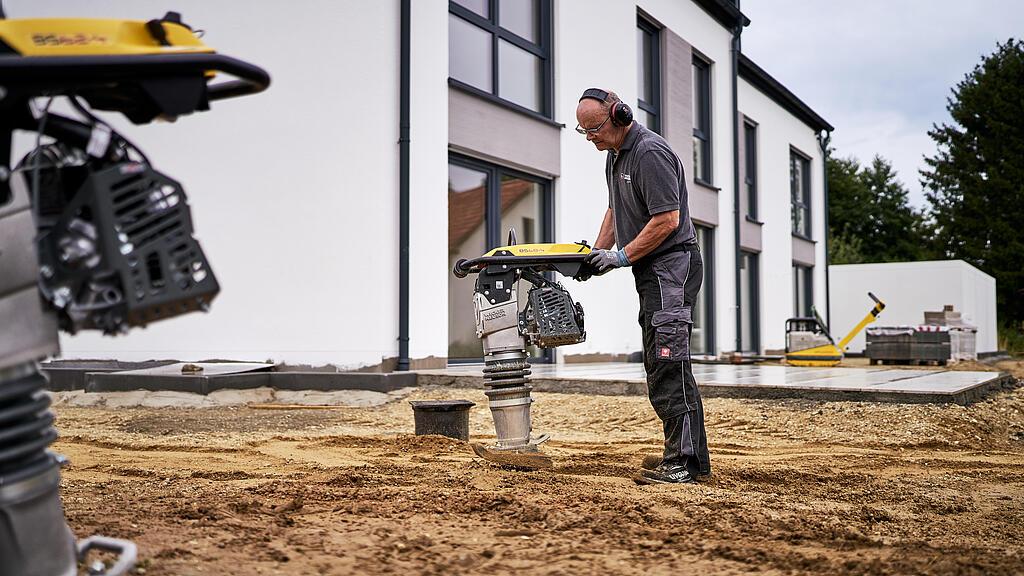 Worker compacts the soil in front of a residential building with a vibratory rammer.